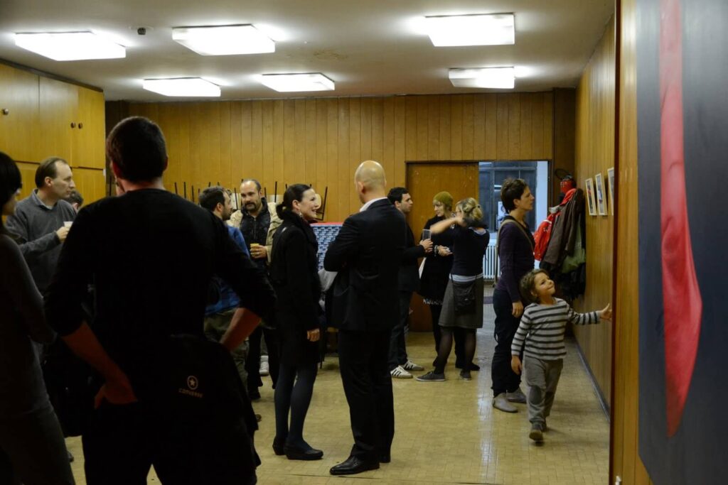 Група људи се дружи у соби обложеној дрветом, неки посматрају уметност на зиду. Мало дете у пругастој одећи се пружа да додирне урамљену слику, што одражава опуштену и живу атмосферу овог окупљања Контекст колектива.