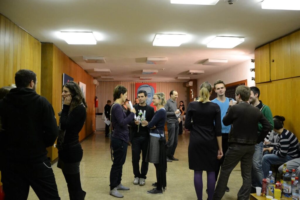 Група људи стоји и разговара у малим групама у јарко осветљеној просторији са дрвеним зидовима, делећи пића и идеје. Столови са флашама су поређани са десне стране, док велики постер на супротном зиду истиче уметност и солидарност.