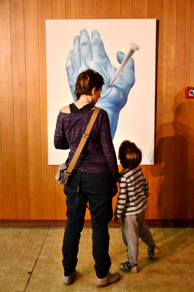 Жена и дете стоје окренути ка слици велике плаве руке прободене ексером, изложеној на зиду обложеном дрвеним панелима. Сцена евоцира уметност и солидарност, а женска торба преко рамена и дечја пругаста одећа додају дубину.