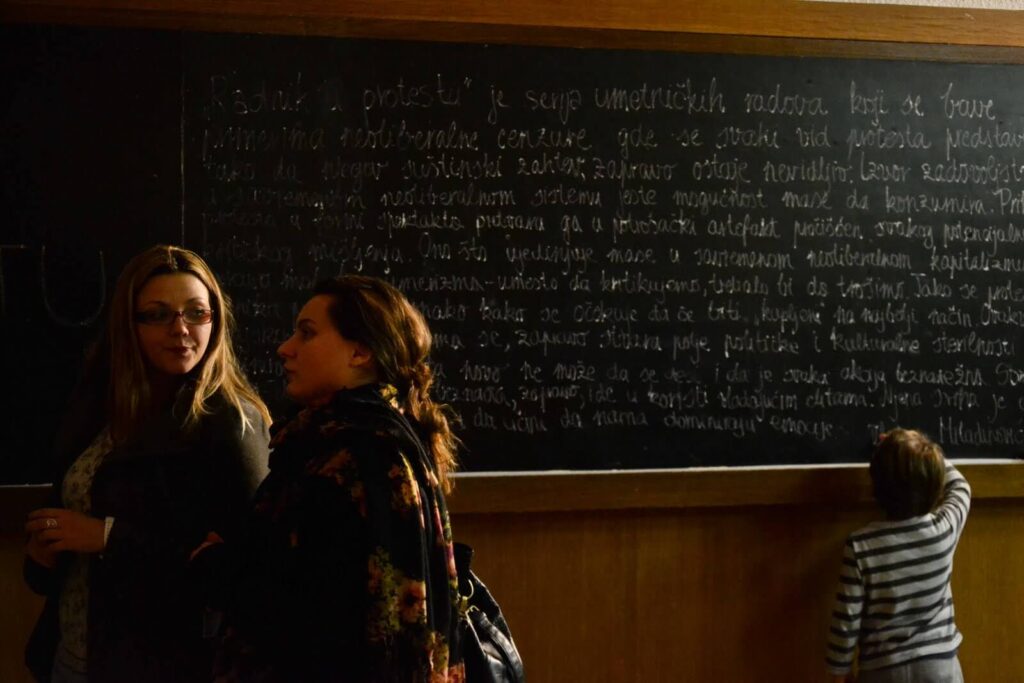 Две жене стоје и разговарају испред велике табле прекривене рукописом писаним текстом, док мало дете у пругастој мајици гледа у таблу. Сцена, пуна топле светлости, одражава дух уметности и активизма.