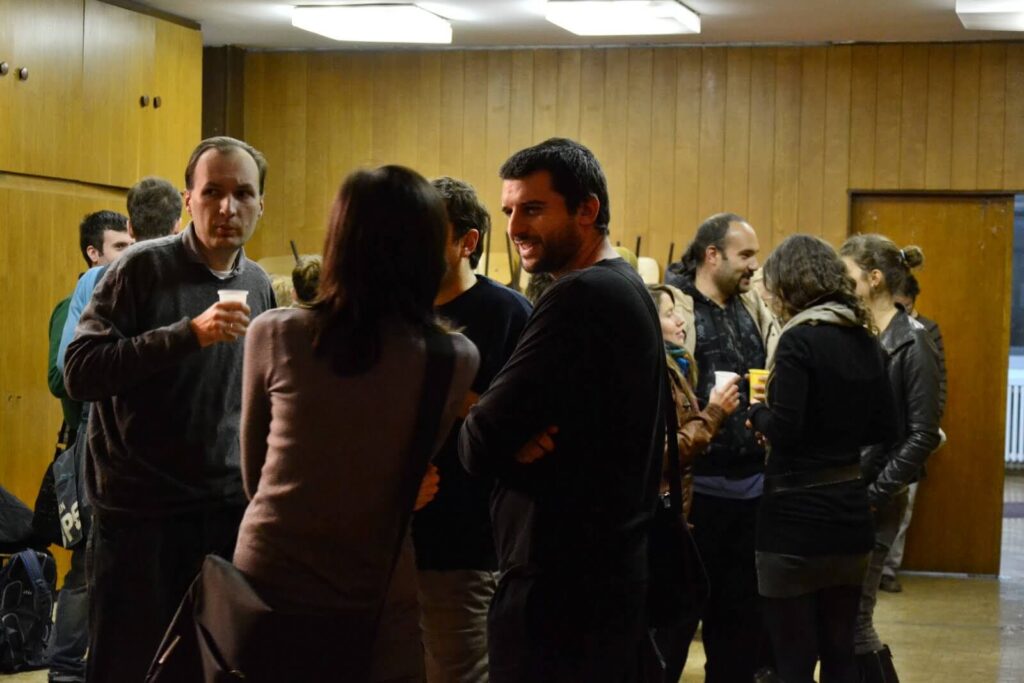 Група људи стоји и разговара у просторији обложеној дрветом, неки држе пића и носе лежерну одећу. Атмосфера делује друштвено и неформално, одражавајући уметност и солидарност унутар колективног окружења.