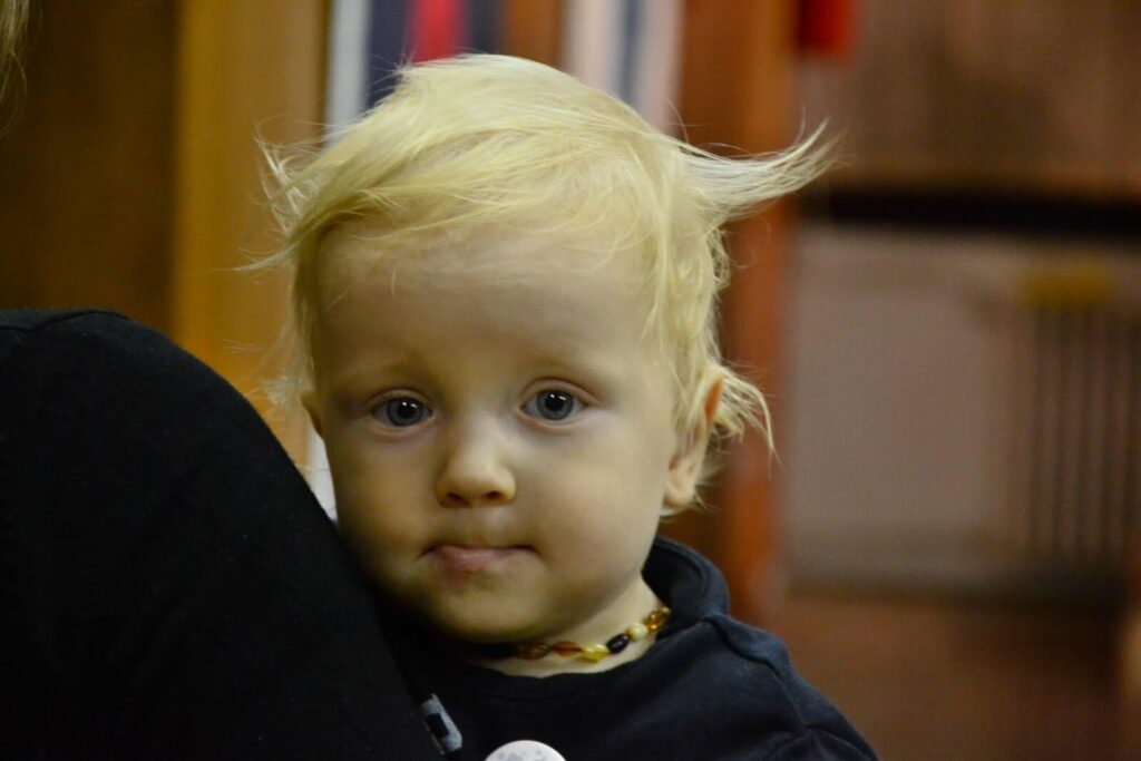 Мало дете са плавом косом и плавим очима гледа у камеру док се наслања на раме одрасле особе, носећи огрлицу од перли и тамну кошуљу. Сцена, благо замућена у позадини, одражава дух уметности и суптилне солидарности.