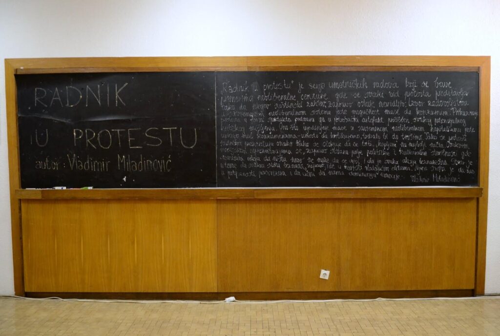 Велика табла са рукописним текстом на српском језику, укључујући наслов РАДНИК У ПРОТЕСТУ и име аутора Владимира Младеновића, постављена је изнад дрвених панела, евоцирајући уметност и солидарност унутар учионице или предаваонице.