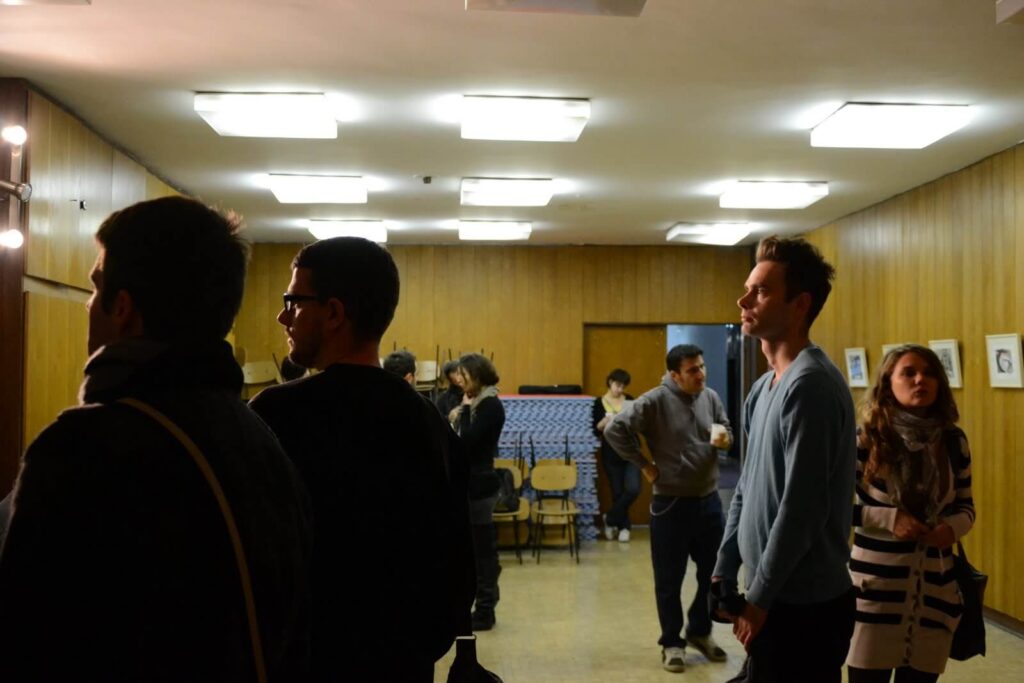 Група људи стоји и хода у просторији обложеној дрветом и са јарким плафонским светлима. Неки виде колективну уметност на зидовима, док други разговарају или се крећу, одражавајући дух солидарности и активизма.