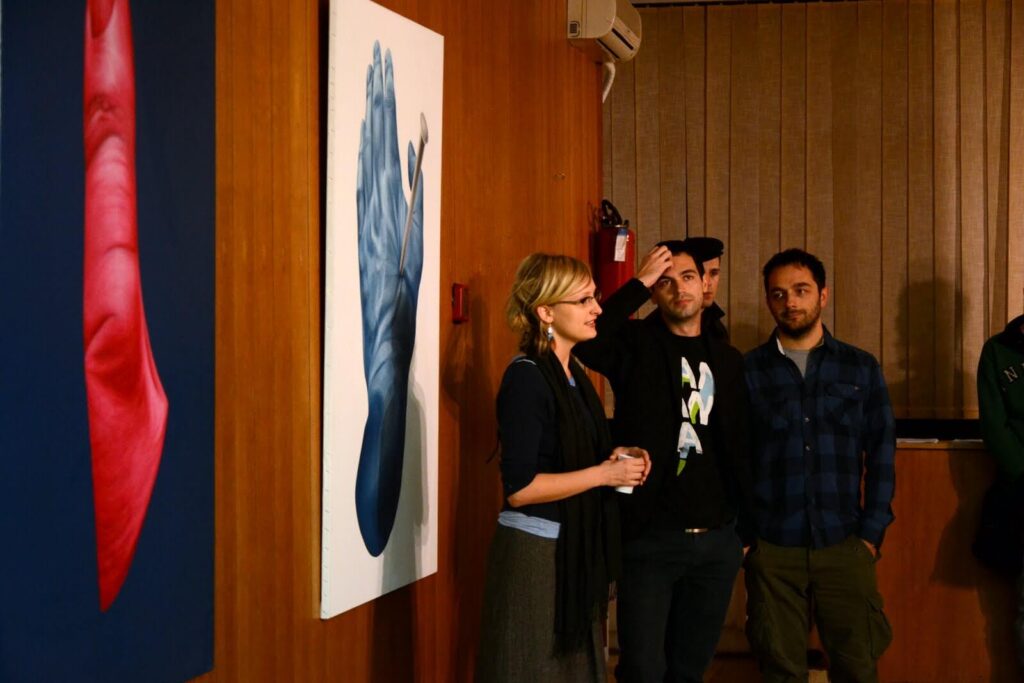 Жена из Контекст колектива говори пред двојицом мушкараца у просторији са дрвеним зидовима, близу апстрактног уметничког дела које приказује плаву руку и црвени облик. Сцена одражава уметност, солидарност и активизам, док други примећују у позадини.