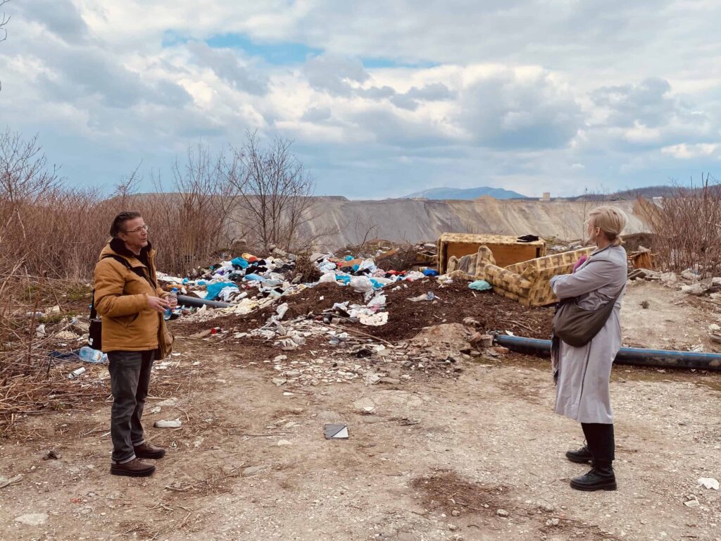Двоје људи стоје на земљаном путу окренути једна према другој близу гомиле смећа и отпада, са одбаченим софама и смећем разбацаним около. Огољено жбуње и планине виде се под облачним небом у позадини.