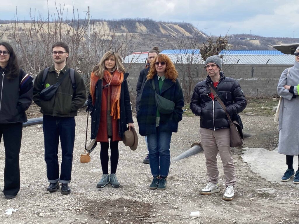 Група од пет одраслих стоји напољу на каменитој површини, обучена у зимску одећу. Иза њих су голо дрвеће, индустријске зграде и брдовити пејзаж под облачним небом.