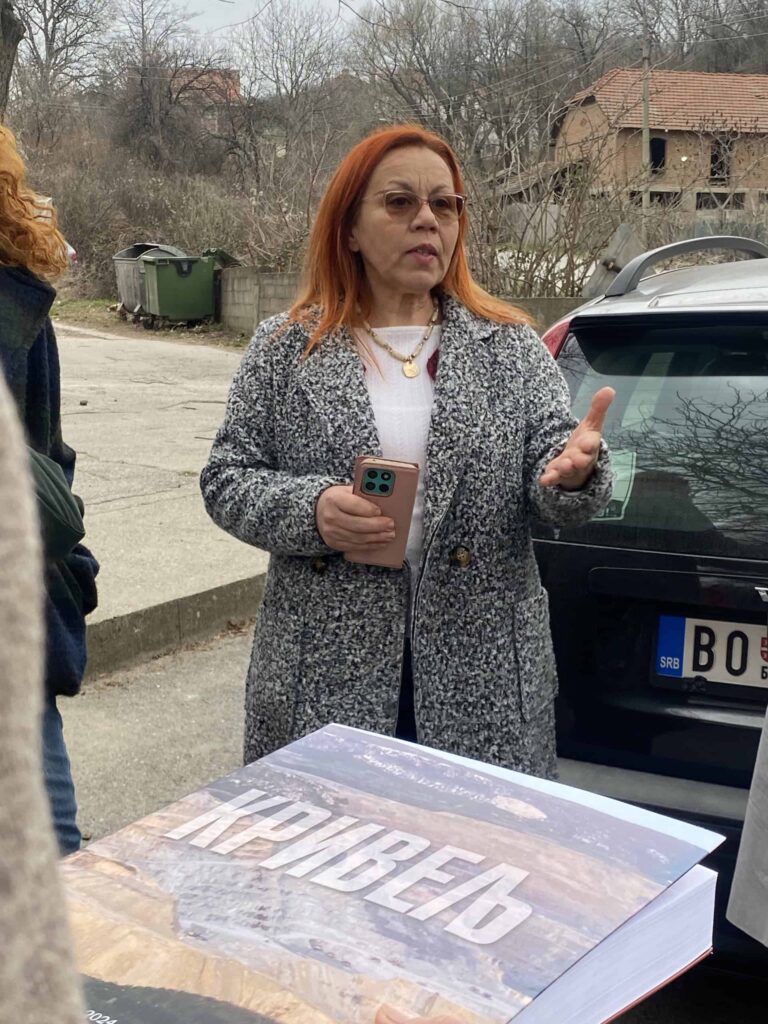 Жена са црвеном косом, са наочарима и сивом капутом, разговара напољу док држи паметни телефон. Испред ње је велика отворена књига са ћириличним текстом. У позадини се виде куће, аутомобил и дрвеће.