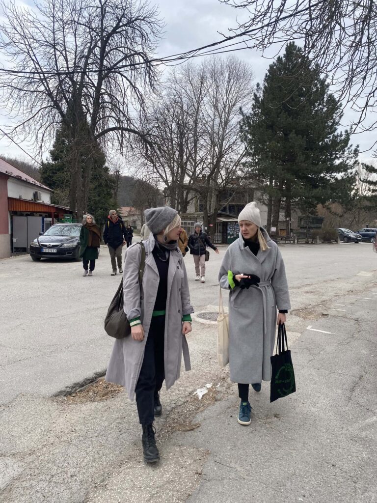 Две жене у сивим капутима и капама шетају и разговарају на паркингу по облачном дану, носећи торбе. У позадини се види још неколико људи и паркираних аутомобила, а у близини се налазе голо дрвеће и зграде.